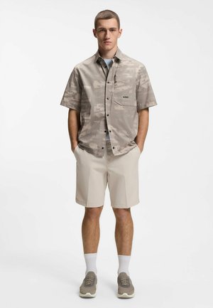 Young man stands with hands in pockets wearing beige patterned short-sleeve shirt, light beige shorts, white socks, and gray sneakers against white background.