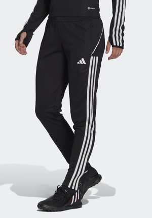 Personne portant un pantalon de sport Adidas noir avec des bandes blanches et des chaussures de sport noires avec des détails bleus et blancs, debout avec les jambes croisées.