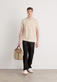 T-shirt beige à manches courtes, pantalon noir texturé et baskets blanches. Portant un sac beige avec des poignées marron. Design simple, look décontracté.