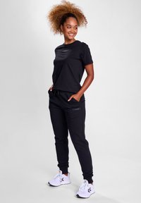 Schwarzes Baumwoll-T-Shirt mit kurzen Ärmeln und Logo auf der Brust, kombiniert mit schwarzen Jogginghosen mit Taschen. Weiße Sportschuhe.