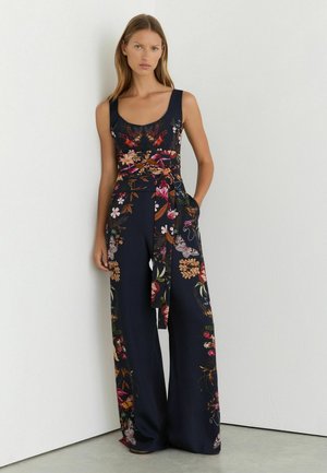 Frau trägt einen ärmellosen marineblauen Jumpsuit mit bunten Blumenmustern und einem gebundenen Gürtel, steht an einer weißen Wand.