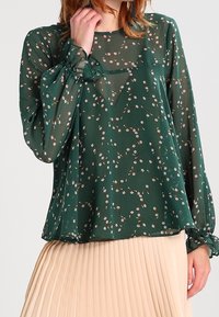 Blouse verte à motif floral avec tissu transparent, manches longues et poignets froncés. Elle présente une coupe ample et de petits motifs de fleurs roses partout.