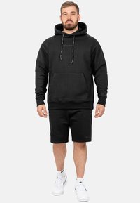 Felpa nera con cappuccio e lacci, tasca frontale e dettagli con logo, abbinata a pantaloni corti coordinati. Modello che indossa sneaker bianche.
