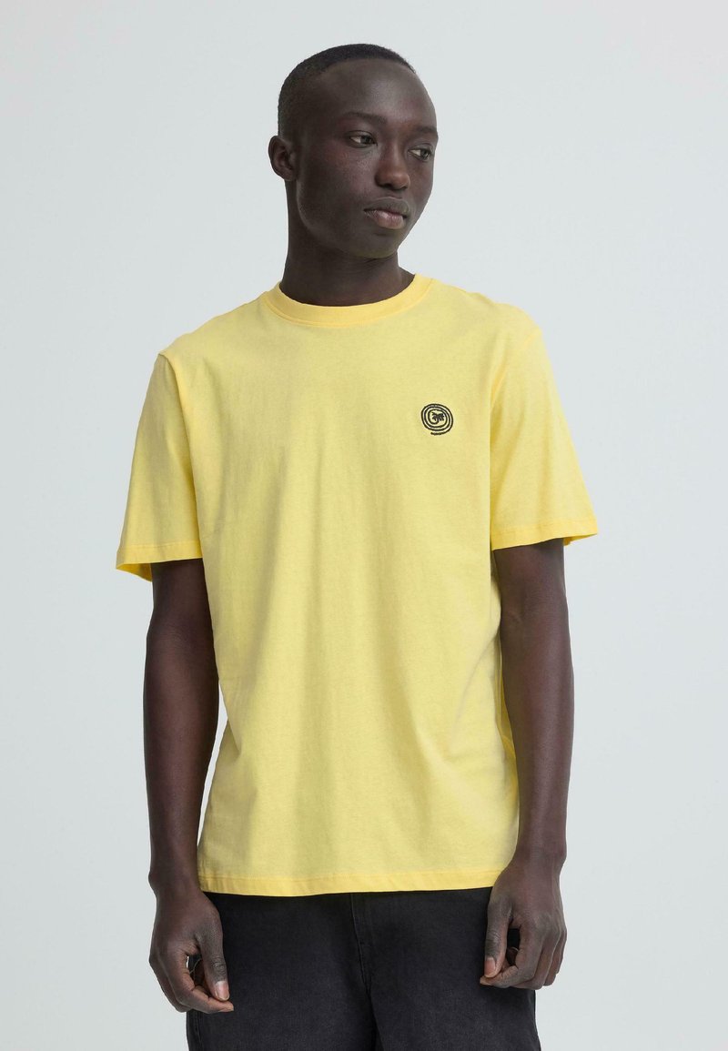 Jeune homme portant un t-shirt jaune uni avec un petit logo circulaire noir sur la poitrine, debout devant un fond gris clair.