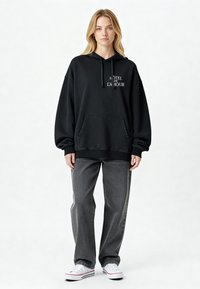 Fekete kapucnis pulóver, elülső zsebbel és fehér szöveggel, amelyen az áll: "HOTEL DE L'AMOUR", szürke, laza szabású farmernadrággal és fehér sportcipőkkel párosítva.