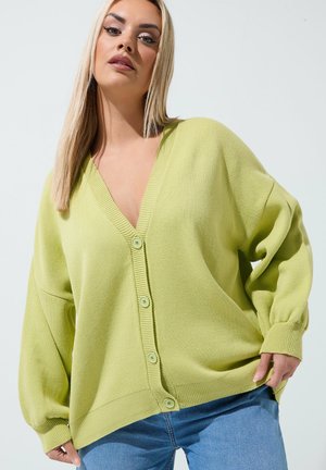 Donna bionda che indossa un cardigan verde chiaro oversize con bottoni e jeans blu, che tiene l'orlo del cardigan, in piedi davanti a uno sfondo neutro.