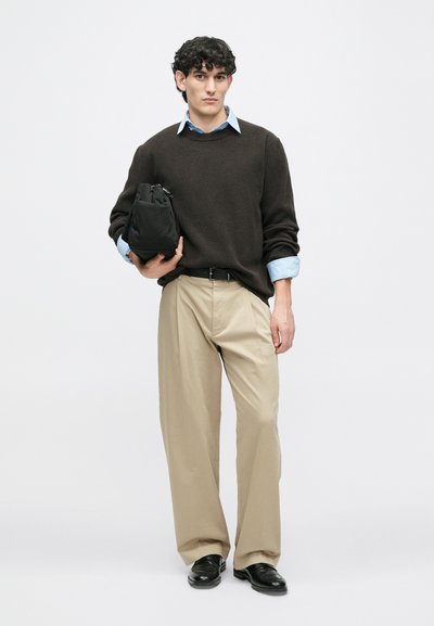 Rudas megztinis, šviesiai mėlynas marškinėlių coloras, smėlio spalvos plačių kojų kelnės, juodas diržas, laikantis juodą rankinę, stovintis ant vienodos fono.