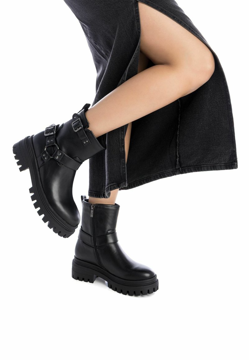 Botas de cuero negras hasta el tobillo con suelas gruesas, con acentos de doble hebilla y cremalleras, combinadas con una falda negra de gran abertura.