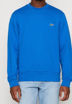 Man draagt een effen felblauwe trui met ronde hals en een klein groen krokodillenlogo op de borst en lichtblauwe spijkerbroek.