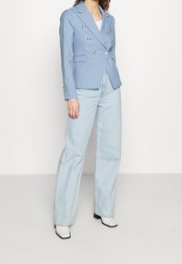 Ljusblå dubbelknäppt kavaj med silverknappar, två framfickor och textilstruktur, kombinerat med ljusa denimjeans och vita anklarstövlar.