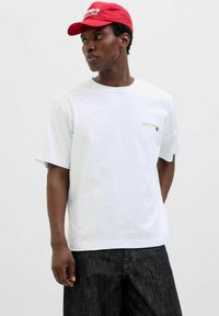 Hvid bomulds t-shirt med kortærmer og et guld "ORIGINALS" logo på brystet, parret med mørk denim jeans og en rød kasket.