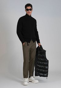 Un pull zippé noir côtelé, un pantalon vert olive, tenant un gilet matelassé noir brillant. Les chaussures sont blanches avec des accents foncés.