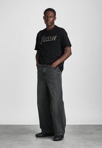 T-shirt preta de manga curta com "VERSACE" escrito em fonte metálica, combinada com jeans cinza escuro de corte largo e sapatos pretos.