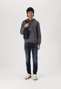 Felpa grigia con zip, jeans blu scuro e sneakers bianche con suole marroni. Una piccola borsa a tracolla nera con cerniera è indossata sulla spalla.