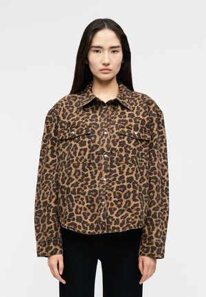 Νεαρή γυναίκα με μακριά σκούρα μαλλιά, ντυμένη με καφέ και μαύρο πουκάμισο με λεοπάρ σχέδιο και μαύρο παντελόνι, στέκεται μπροστά σε απλό φόντο.