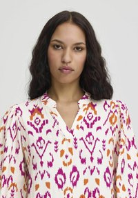Flowy, patterned shirt in off-white with vibrant purple and orange abstract designs. Features a notch collar and puffed sleeves.