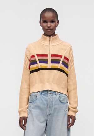 Persona che indossa un maglione beige lavorato a maglia con righe orizzontali rosse, verdi, blu, gialle e nere e jeans azzurri usurati.