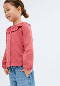 Nuori tyttö iloisen ilmeen kanssa, päällään vaaleanpunainen, teksturoitu paita röyhelökauluksella ja siniset farkut, seisoo vaaleaa taustaa vasten.