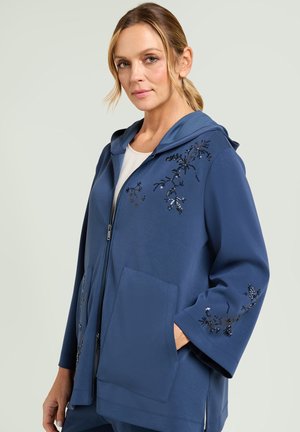 Mujer con sudadera azul con cremallera, bordado de lentejuelas florales negras en el hombro y la manga, manos en los bolsillos delanteros, fondo claro.