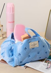 Bolsa de peluche azul claro con lazos bordados, que contiene un cepillo de pelo rosa, una botella de champú rosa y otros artículos de tocador sobre una mesa.