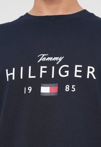 Námořnická modrá košile s bílým nápisem "Tommy HILFIGER", rokem "1985" a malým červeným, bílým a modrým logem vlajky Tommy Hilfiger.