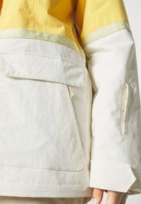 Een anorak met een gele bovenkant en een witte onderkant, voorzien van een grote voorkenn Pocket, zijritsen en een patroonrand langs de mouwen.