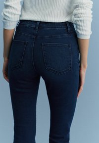 Jeans din denim de un albastru închis, cu un design slim, având două buzunare la spate și un talie înaltă. Materialul are o textură netedă.