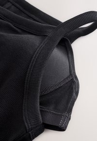 Gros plan sur un tissu noir avec bord côtelé et coutures, montrant une partie d'une bretelle ou d'un col de vêtement plié sur un fond clair.