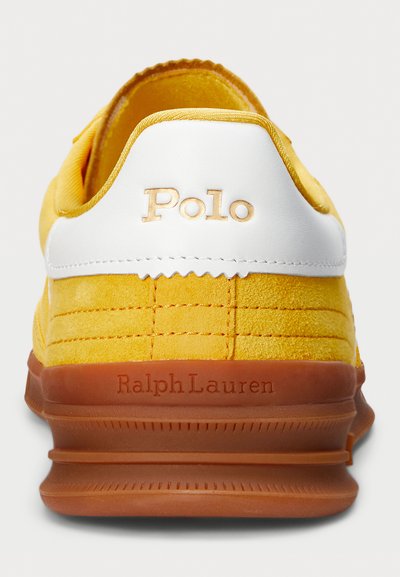Baskets en daim jaune avec un accent en cuir blanc, comportant un logo « Polo » embossé, une semelle en caoutchouc marron et un détail texturé au niveau du talon.