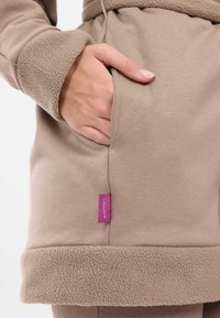 Felpa beige con colletto e polsini foderati in morbido pile, tasca laterale e un'etichetta viola prominente sulla cucitura.