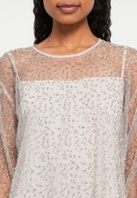 Egy nő áttetsző, világos színű felsőt visel, amelyet kis, csillogó flitterek díszítenek, egy hasonló színű camisole fölött.