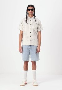 Camicia a maniche corte di un beige chiaro con stampa floreale a contorno, abbinata a pantaloni corti di jeans blu chiaro, calze bianche e sneakers color beige con accenti rossi.
