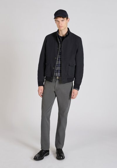 Chaqueta de algodón azul marino con un cuello estructurado y cierre de botón, combinada con una camisa de cuadro oscuro, pantalones grises y zapatos negros.