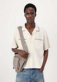 Short-sleeve, cream shirt with black piping, two chest pockets, and a relaxed fit, paired with a gray sling bag featuring front pocket and flap.