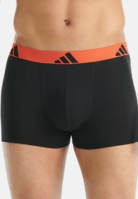 Svarta boxershorts med en tjock orange och svart märkesresår. Tyget verkar mjukt och figurnära, vilket betonar en modern design.