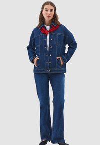 Kvinna klädd i en mörkblå denimjacka och jeans med en vit skjorta och en röd scarf knuten runt halsen, stående med händerna i fickorna.