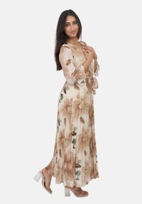 Gemaakte lange jurk met beige en bloemenprint, voorzien van doorschijnende lange mouwen en een strikceintuur; gecombineerd met transparante blokhak sandalen.