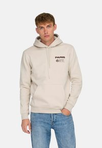 Sudadera beige con un bolsillo delantero, capucha con cordones y logo impreso "PARÍS" en marrón. Combinada con vaqueros azules de corte casual.