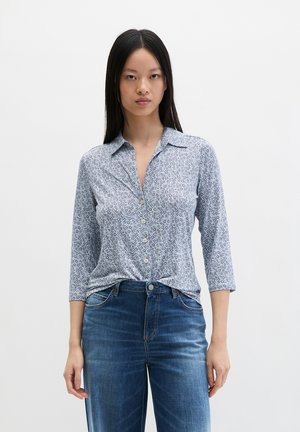 Woman wearing a light blue floral button-up shirt with three-quarter sleeves and blue jeans against a plain white background.