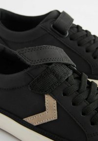 Černé tenisky s suchým zipem, kulatými tkaničkami, béžovým akcentem ve tvaru šípu a texturovanou gumovou podrážkou.