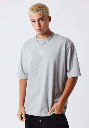 Jeune homme aux cheveux blonds courts portant un t-shirt gris oversize avec un petit logo blanc et un collier chaîne argentée.