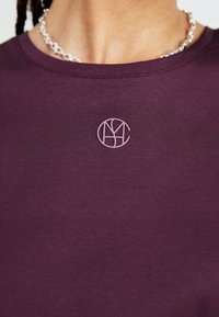 Maroon cotton t-shirt featuring a small lavender-colored embroidered logo on the chest. Smooth texture with a round neckline.