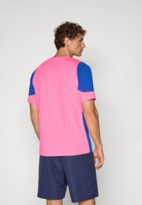 Αθλητικό μπλουζάκι με κοντά μανίκια, με ροζ πλάτη και μπλε μανίκια. Φτιαγμένο από απαλό ύφασμα; συνδυάζεται με σκούρα σορτς. Απλός σχεδιασμός, χωρίς σχέδια.