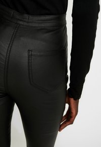 Svart imitation läderbyxor med hög midja, bakficka och en slät yta. Närbild som framhäver sömdetaljer och passform.