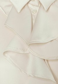Light beige blouse featuring a ruffled collar, soft texture, and a single button closure. The fabric has a smooth and subtle sheen.