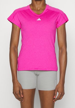 Nainen, jolla on kirkkaan vaaleanpunainen lyhythihainen urheilupaita ja vaaleanharmaat tiukat shortsit, seisoo yksinkertaisen taustan edessä.