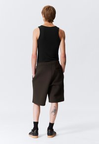 Top negru fără mâneci, asociat cu pantaloni scurți bermați maro, având o textură netedă și buzunare. Tatuaj evidențiat pe partea inferioară a piciorului. Pantofi negri cu talpă de cauciuc.