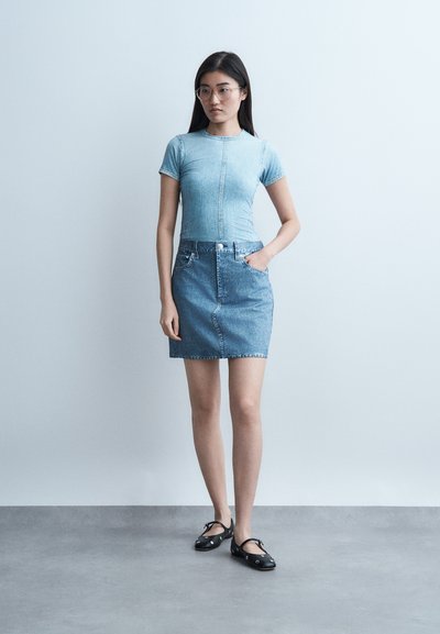 Νεαρή γυναίκα με μακριά σκούρα μαλλιά και γυαλιά, ντυμένη με στενή ανοιχτό μπλε denim μπλούζα και φούστα, που στέκεται μπροστά σε έναν απλό ανοιχτό τοίχο.