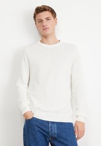 Witte gebreide trui met een ronde hals en ribbels aan de mouwen, gedragen over blauwe denim jeans. Heeft een subtiele logo op de borst.