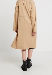 Manteau long beige à coupe droite, manches longues et fente au dos. Associé à des bottines noires, mettant en valeur une texture de tissu lisse.